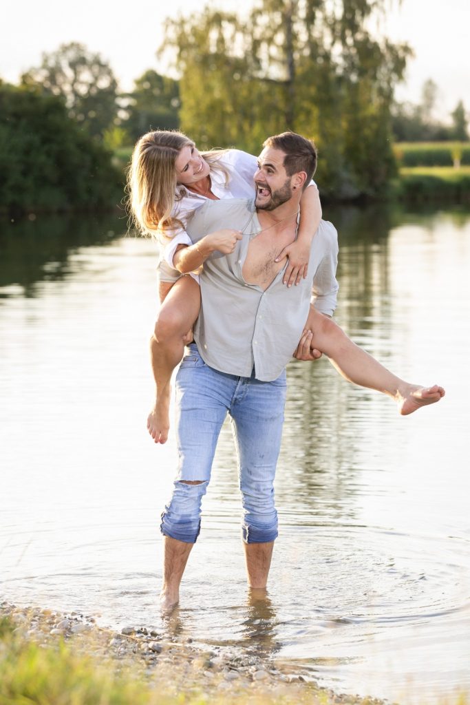 Mann trägt Frau huckepack durchs Wasser, beide lachen